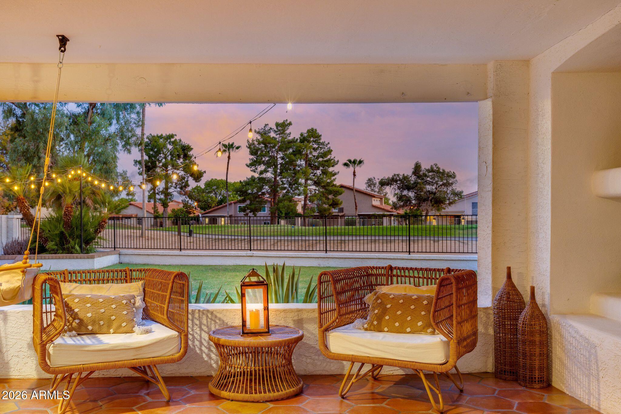 Covered patio with wicker seating and glowing string lights overlooking a fenced lawn at dusk.