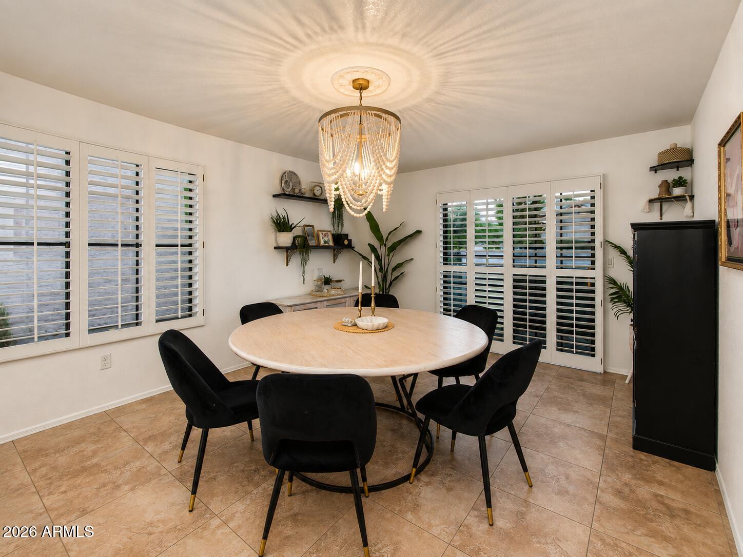 Dining room with a round wood table, black chairs, plantation shutters, and a beaded chandelier.