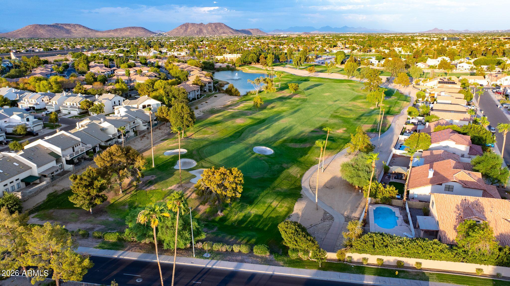 Aerial view of a golf course winding among homes, with a lake and mountains in the distance.