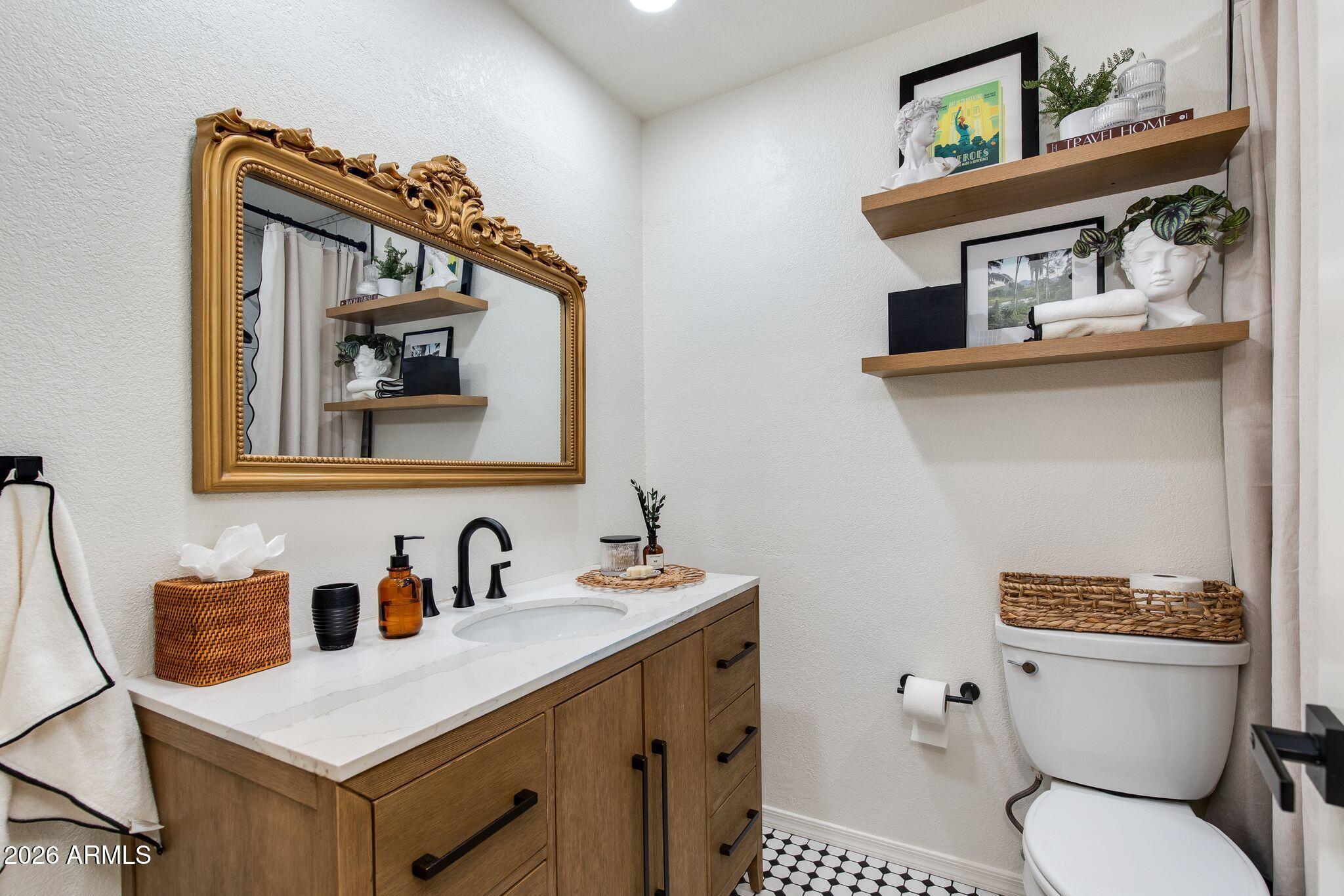 Bathroom with a wood vanity, ornate gold mirror, floating shelves, and black-and-white hexagonal tile floor.