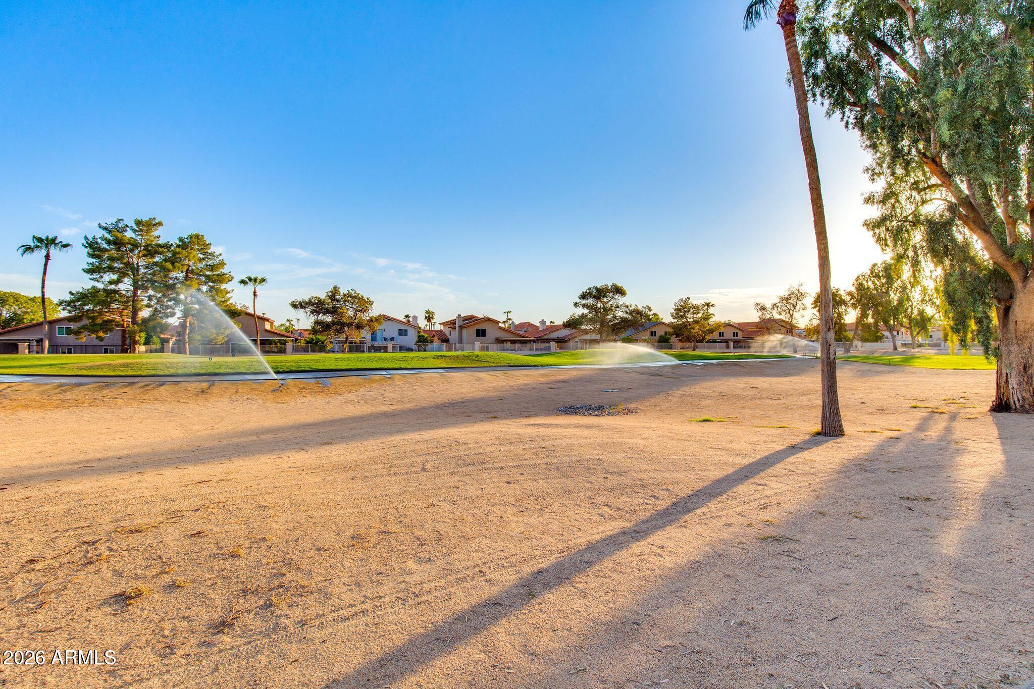 Open park area with sprinklers, mature trees, and homes in warm evening light.