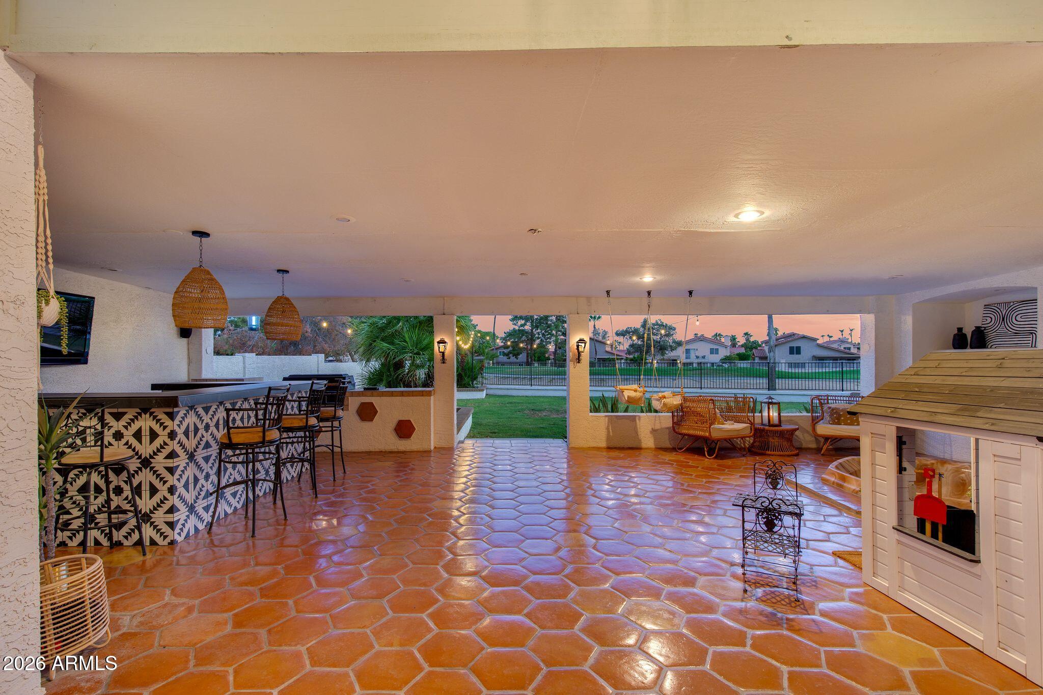 Covered patio with terracotta tile, patterned bar, wicker seating, and hanging chairs at dusk.