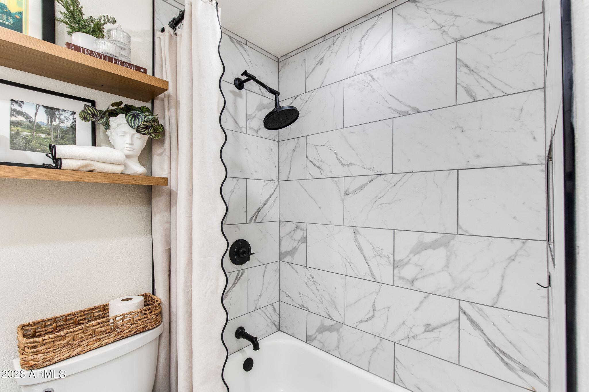 Bathroom with a marble-look tiled tub surround, matte black fixtures, and styled wooden shelves.