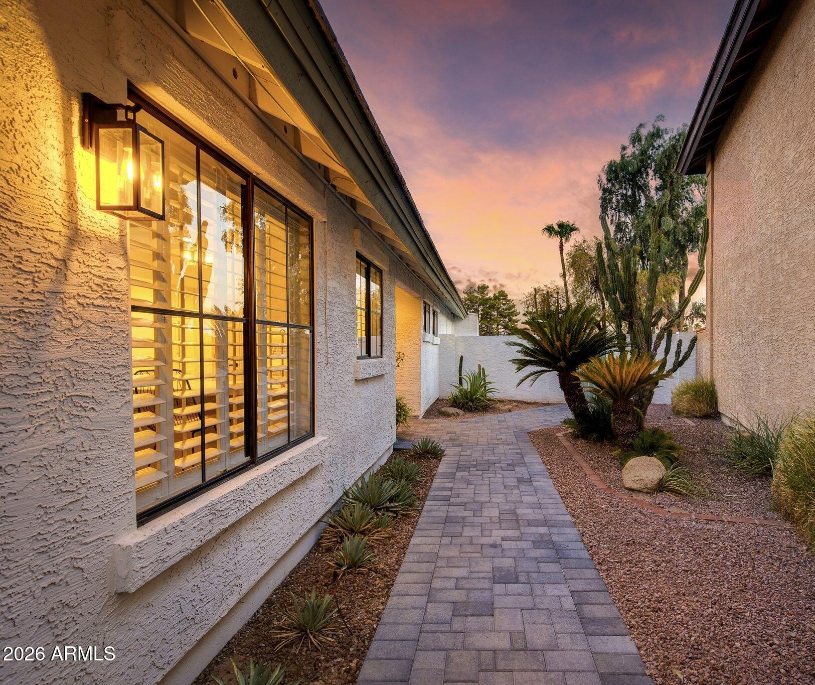 Stucco home with warmly lit windows and a desert garden along a paved walkway at sunset.