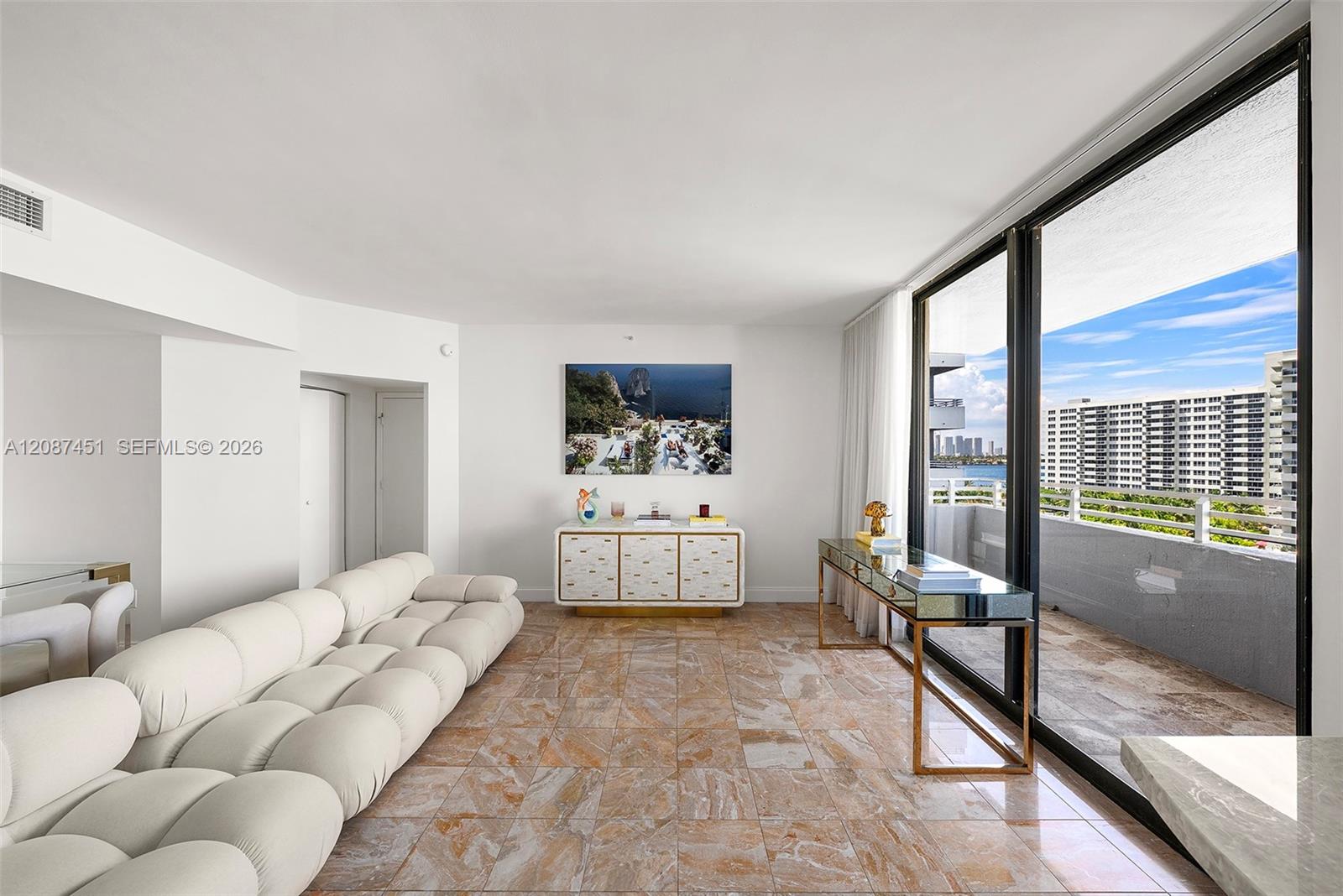 Furnished living room with a white sectional sofa and balcony windows overlooking water and high-rise buildings.