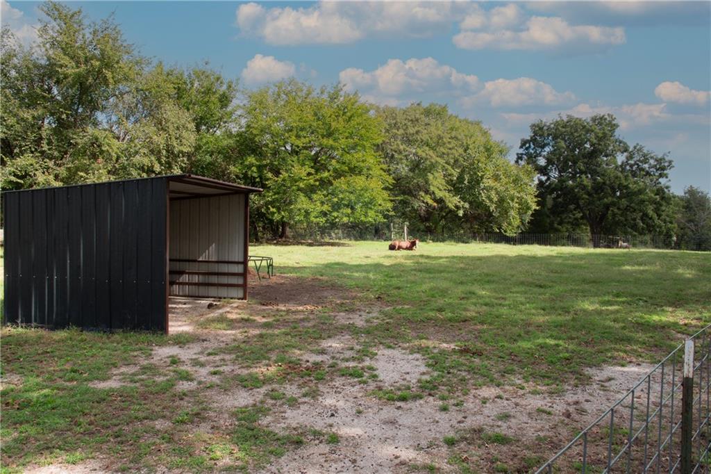 Fenced grassy property with an open-sided black metal shelter and mature trees.