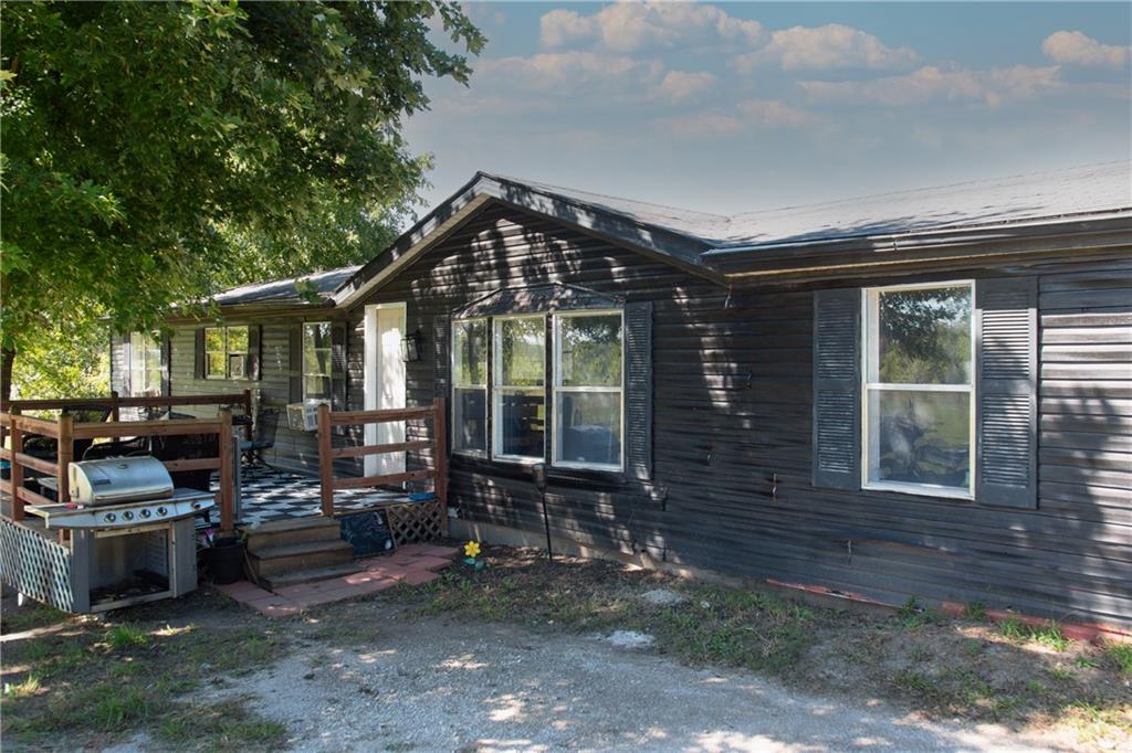 Single-story home with dark siding, raised deck, and outdoor grill beneath leafy trees.