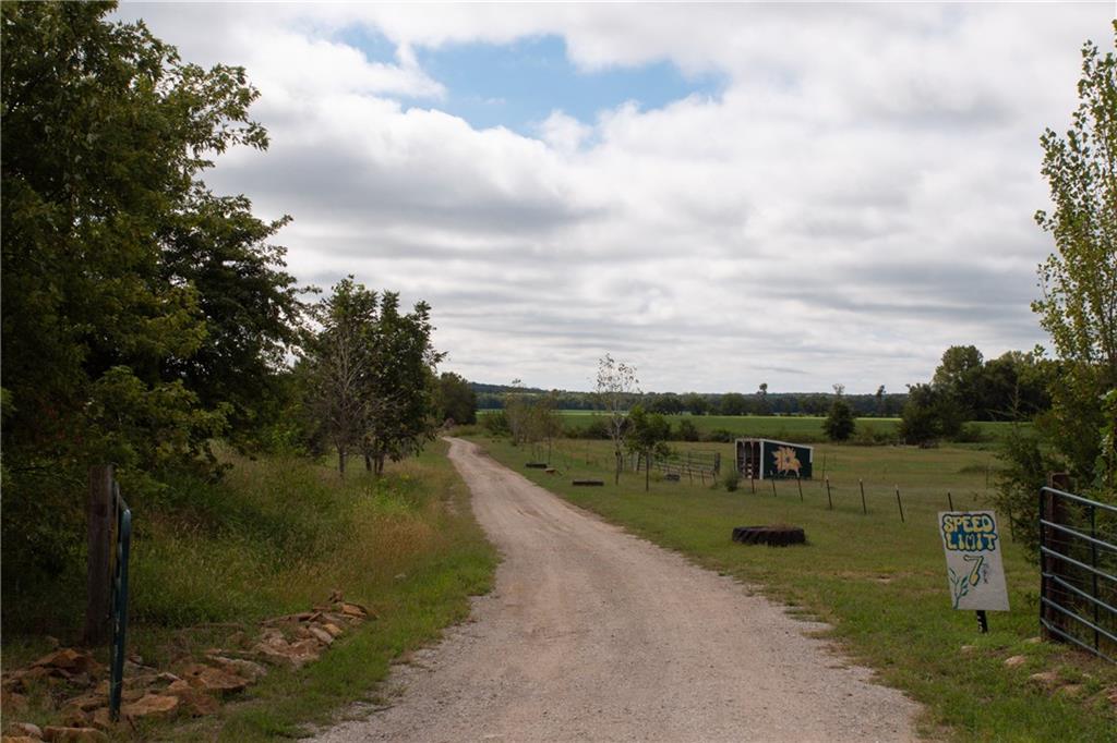 Gravel driveway through a rural property toward open fields, bordered by trees and fencing.