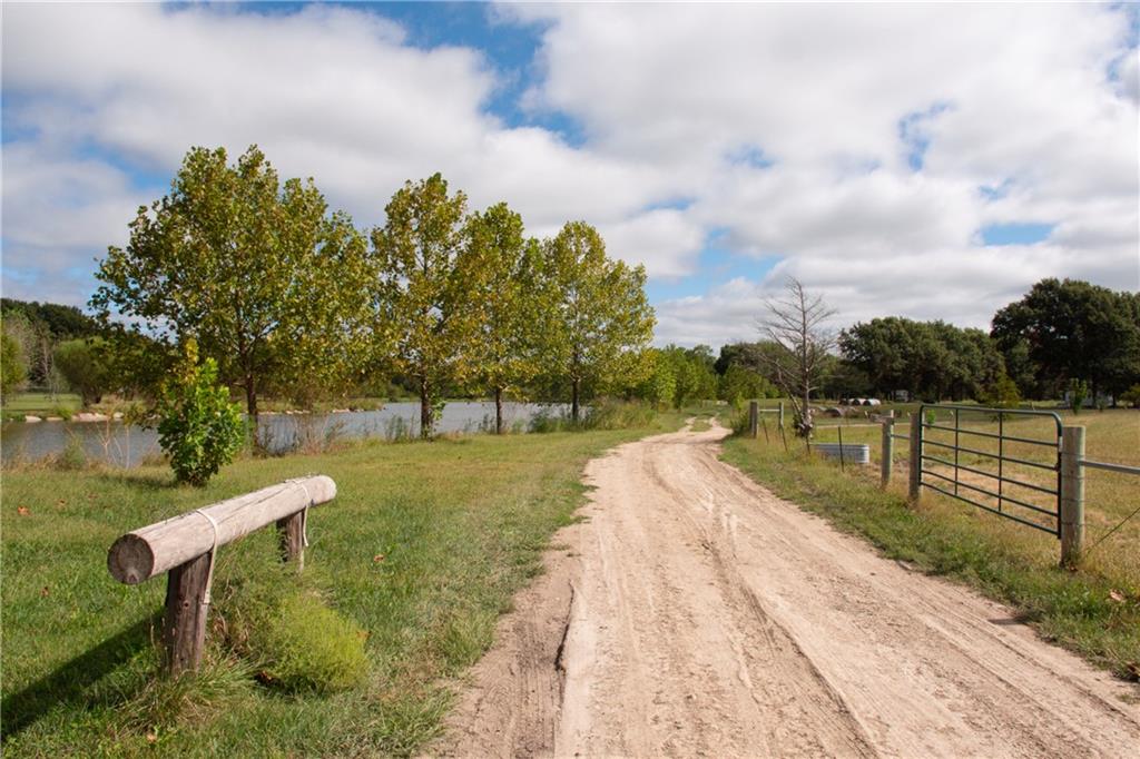 Dirt driveway curves beside a pond toward a metal gate on a rural property.