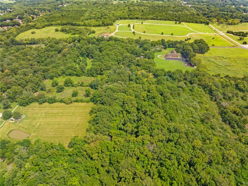 Aerial view of wooded acreage with open fields, a small pond, and a building beside a loop drive.