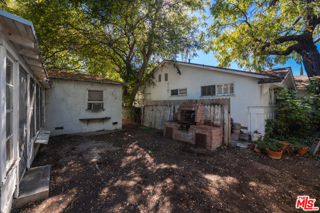 House rear exterior and shaded backyard with a brick barbecue, wooden fence, and mature trees.