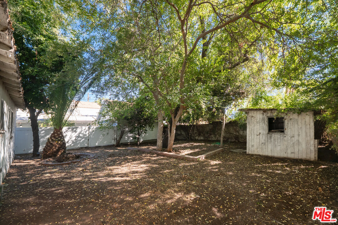 Shaded backyard with mature trees, a small white shed, and a white fence.