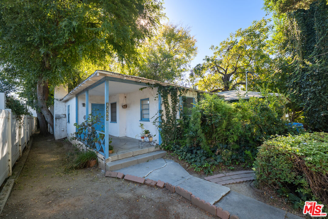 White single-story house with blue-trimmed porch, surrounded by mature trees and dense greenery.