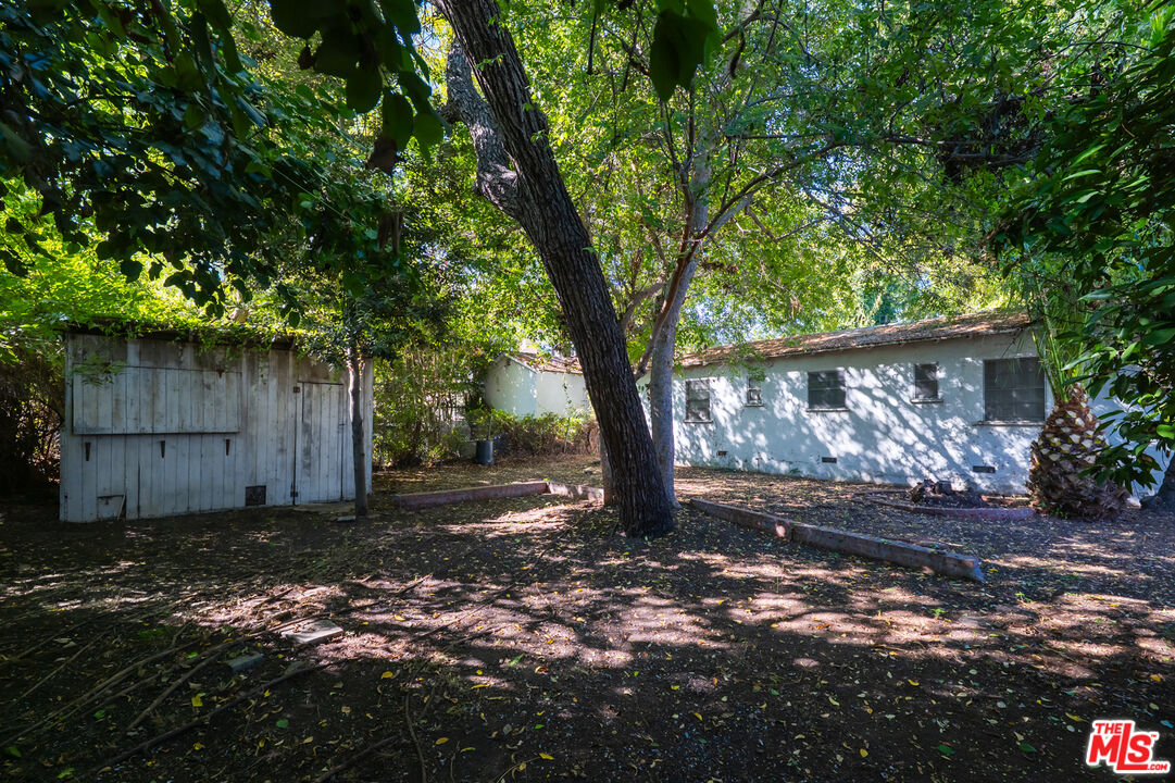 Shaded backyard with mature trees, a wooden shed, and a low white outbuilding.