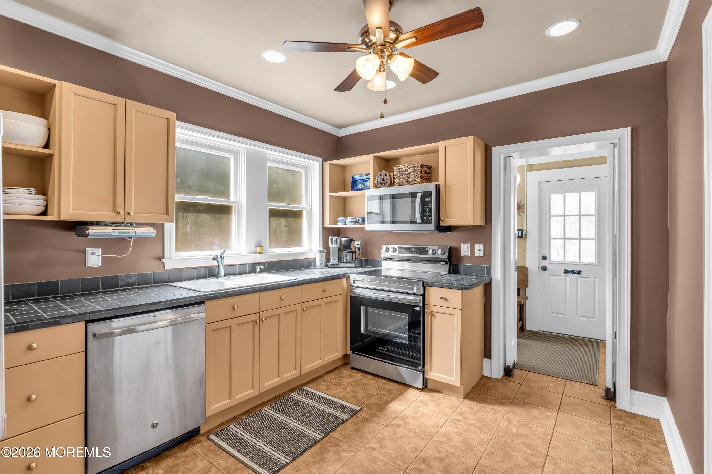 Kitchen with tan cabinets, dark tile counters, stainless steel appliances, and two windows above the sink.
