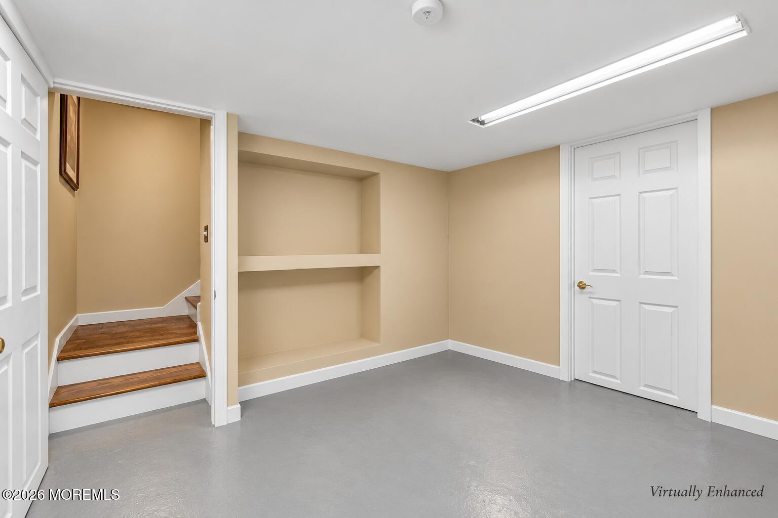 Empty room with built-in shelves, gray flooring, and wood-tread steps to an adjoining area.