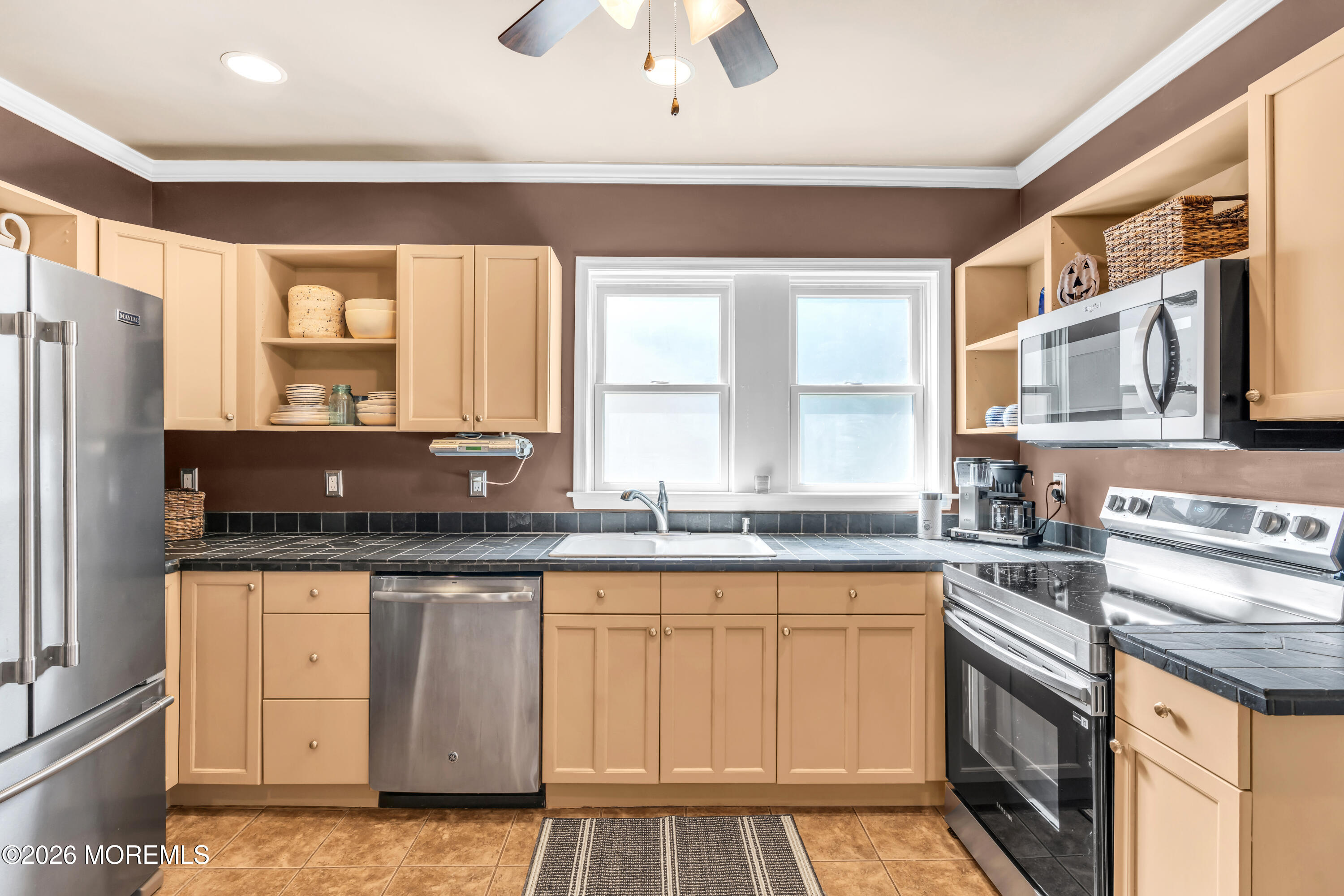 Kitchen with tan cabinets, dark tile counters, a double window, and stainless steel appliances.