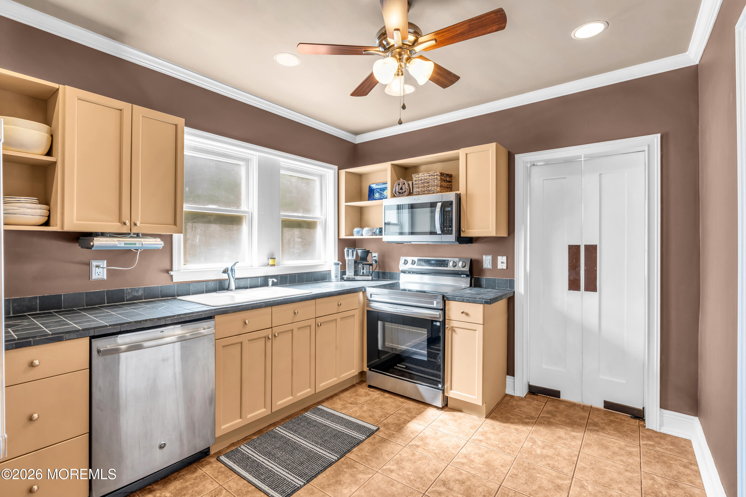 Kitchen with tan cabinetry, dark tile counters, stainless steel appliances, and windows above the sink.