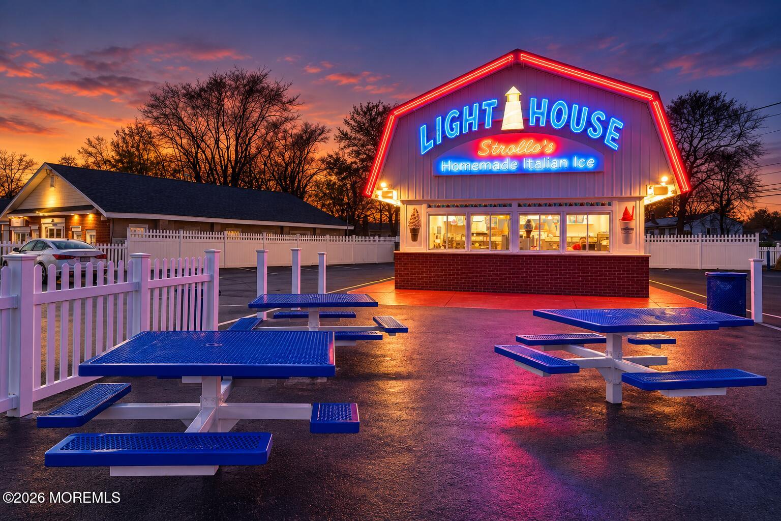Lighthouse Strollo’s Homemade Italian Ice shop glowing at dusk, with blue picnic tables in the foreground.