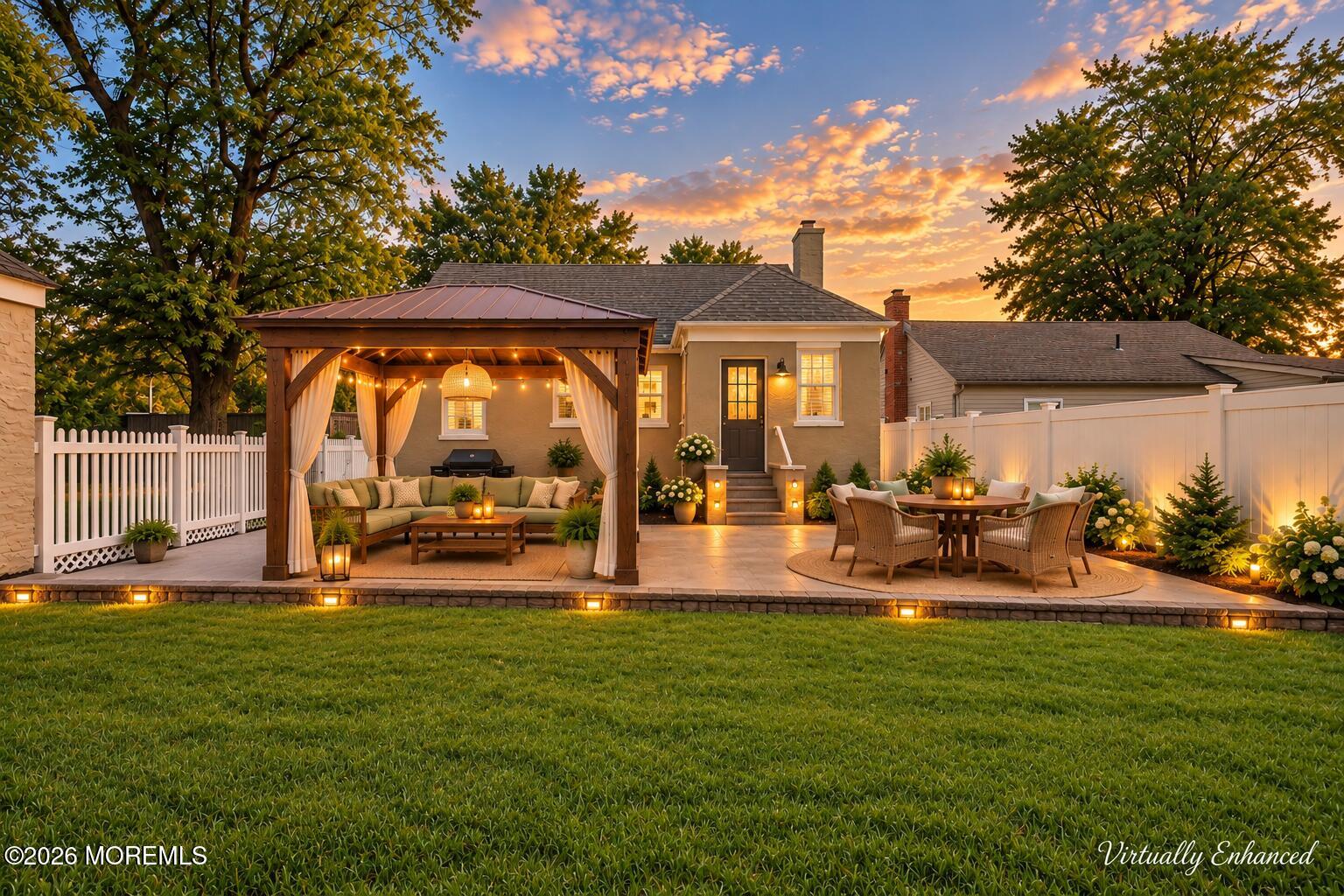 Home’s rear patio with an illuminated pergola, outdoor dining area, and fenced lawn at sunset.
