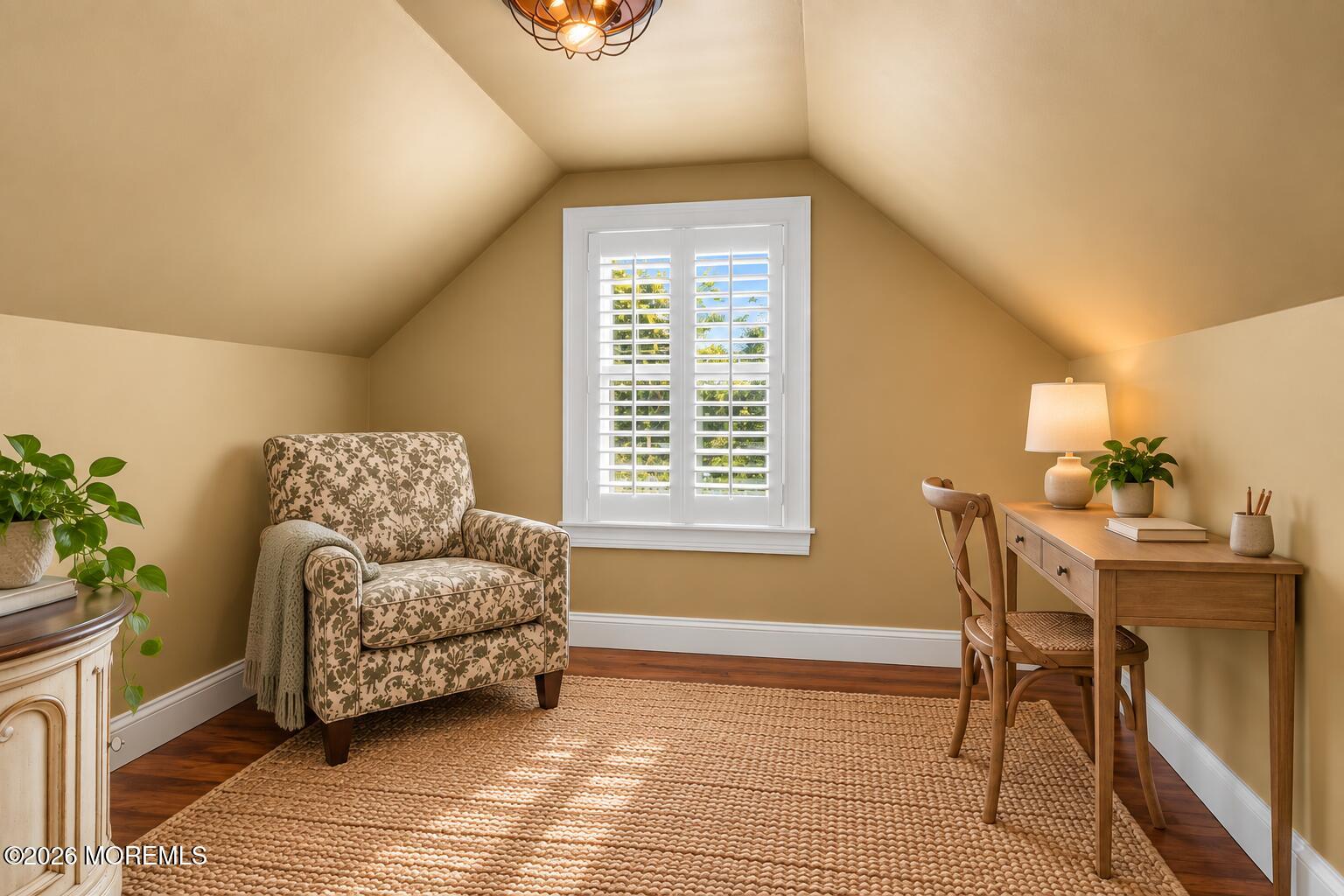 Furnished home office with a patterned armchair, wooden desk, and shuttered window beneath a sloped ceiling.