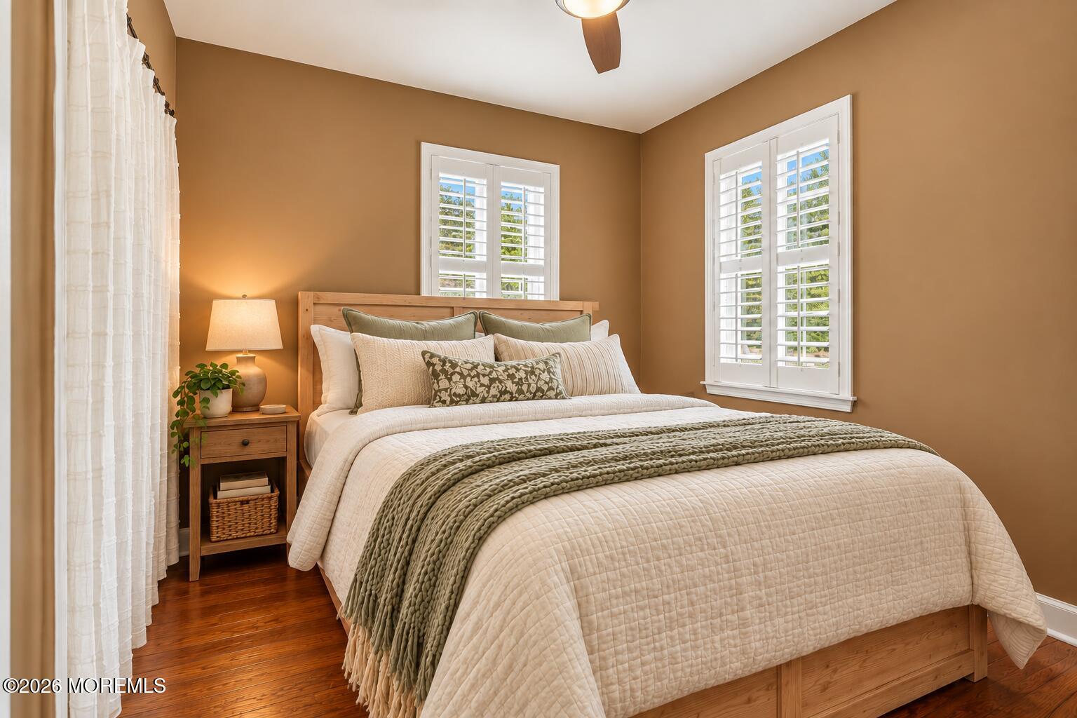 Staged bedroom with a wooden bed, cream bedding, sage accents, and white plantation shutters.