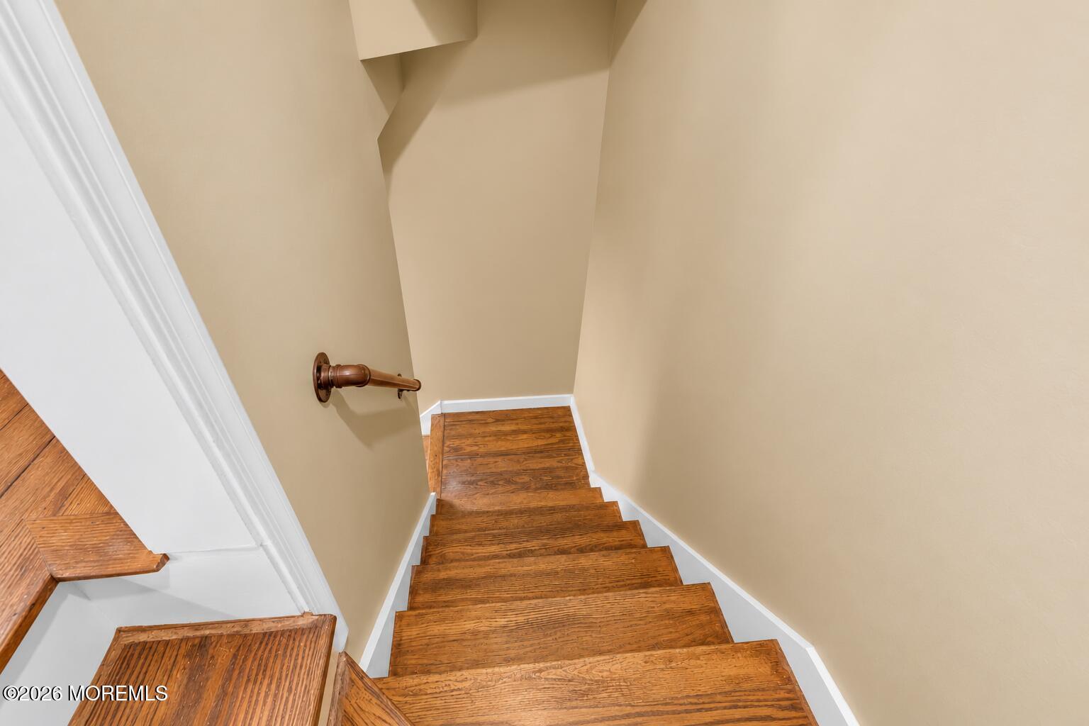 Wooden staircase descending through a beige-walled stairwell with white trim and a wooden handrail.