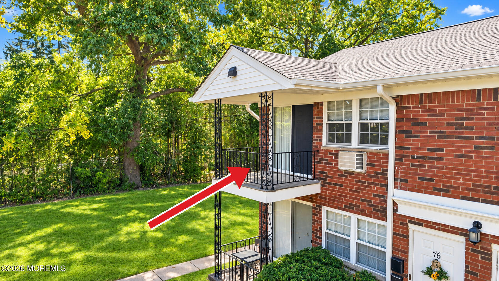 Brick multi-unit building with a raised balcony, decorative iron railings, and a leafy lawn.
