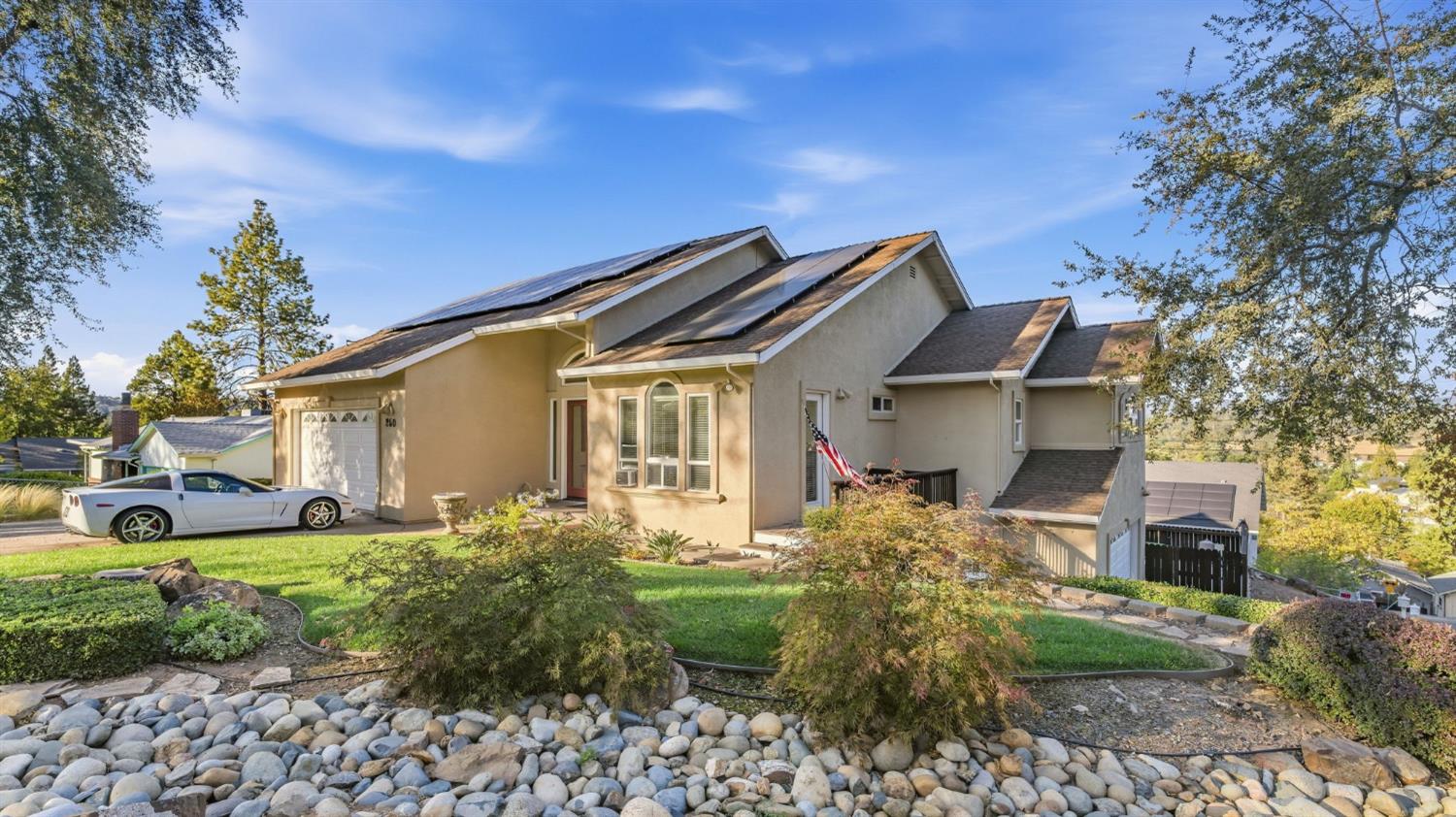 Beige multi-level home with rooftop solar panels, a white sports car, and a landscaped rock garden.