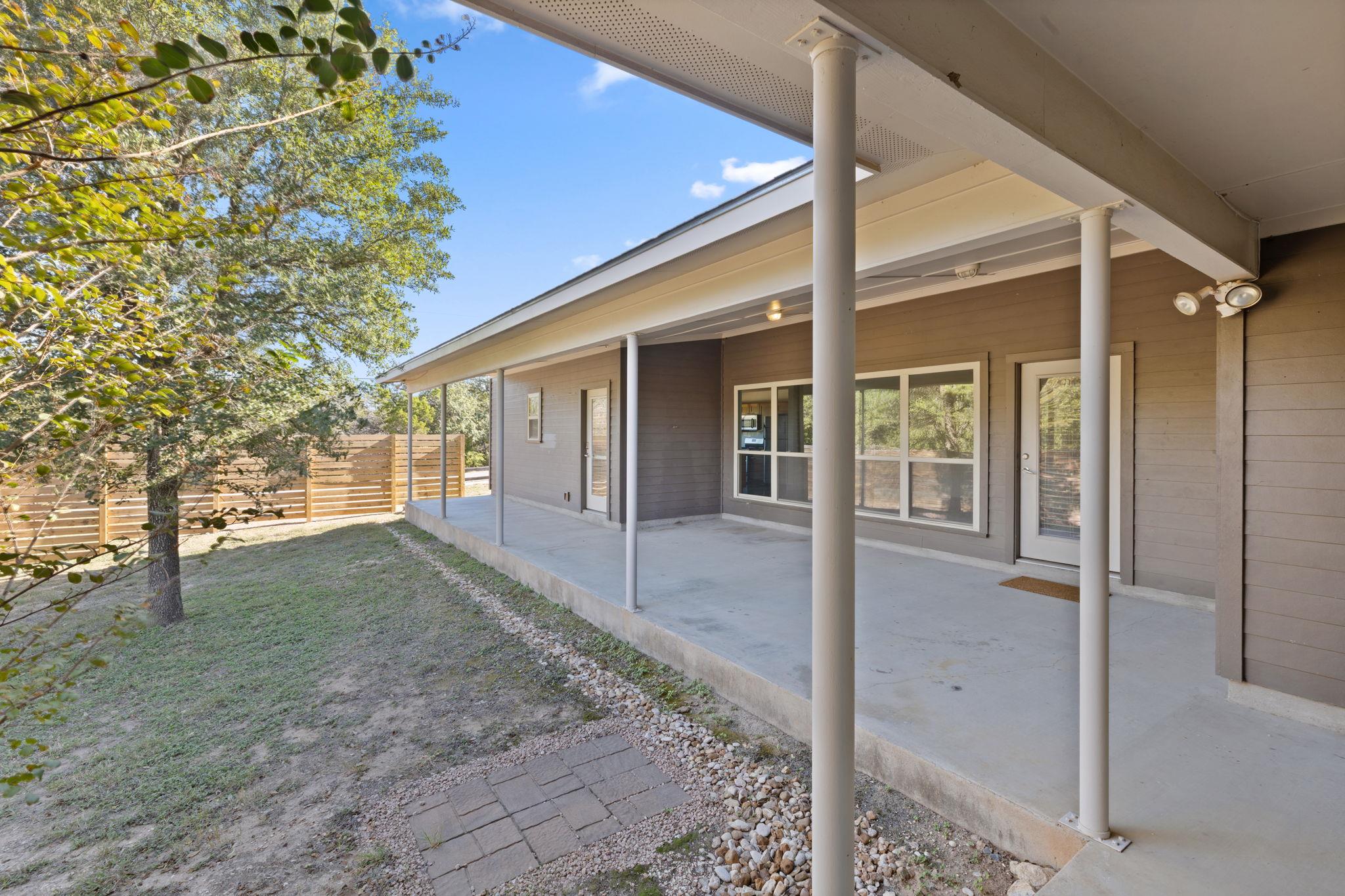 Gray home’s covered back patio with concrete flooring, glass doors, and a fenced, tree-lined yard.