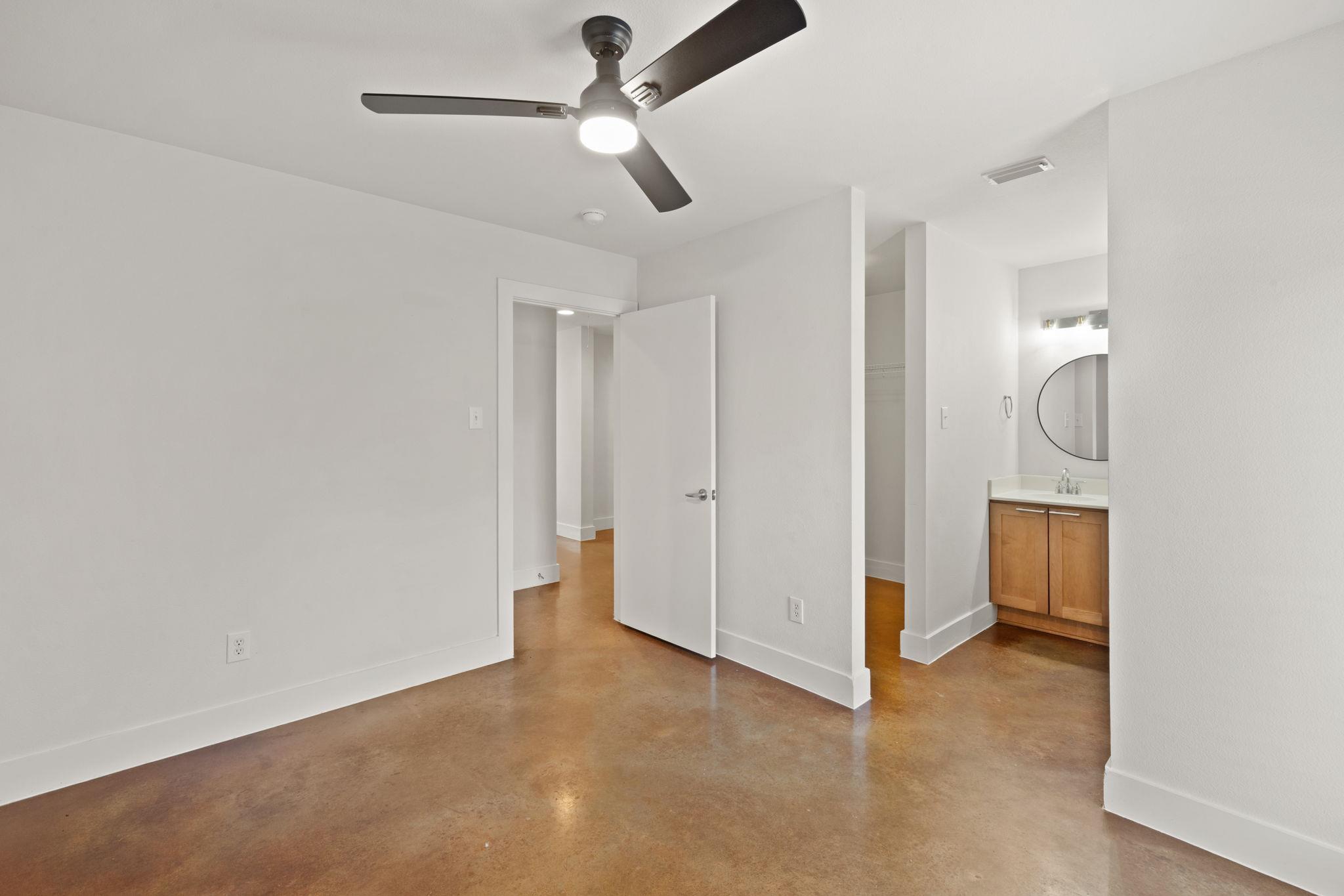 Empty bedroom with polished concrete floors, ceiling fan, and adjoining bathroom vanity.