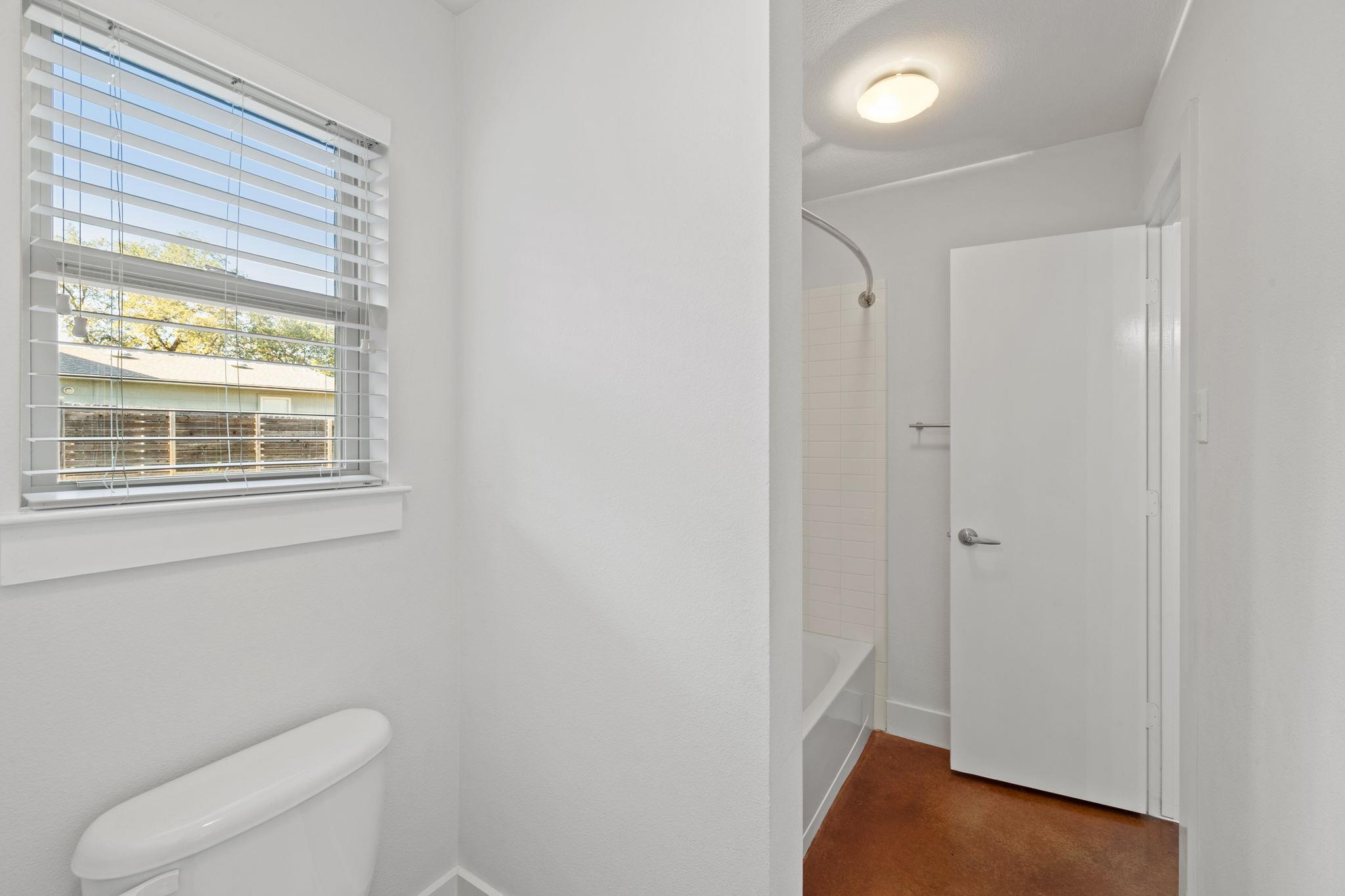 Bathroom with a toilet by a window and a bathtub-shower beyond the open doorway.