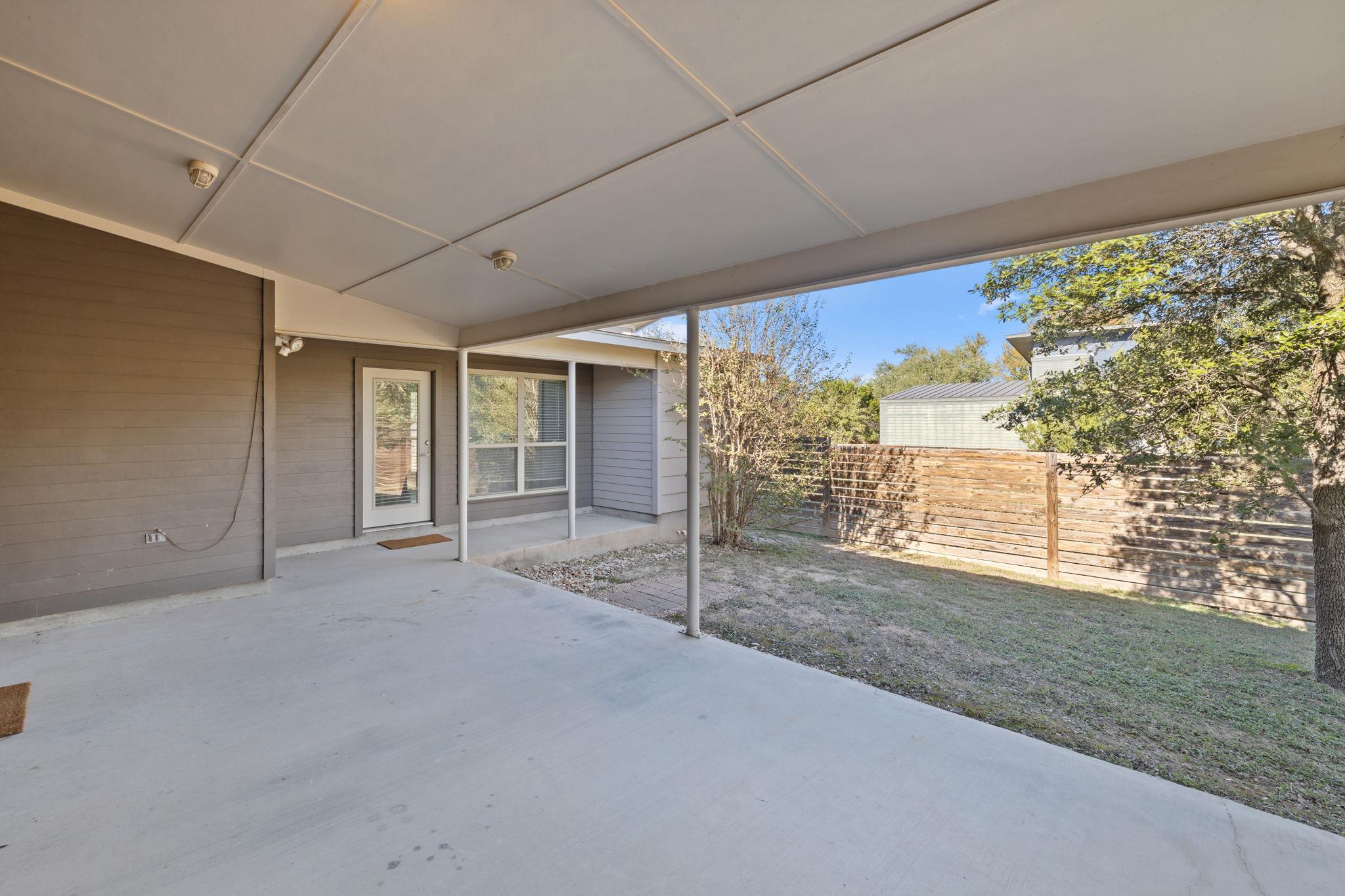 Covered concrete patio beside a gray-sided home, overlooking a fenced backyard with trees.
