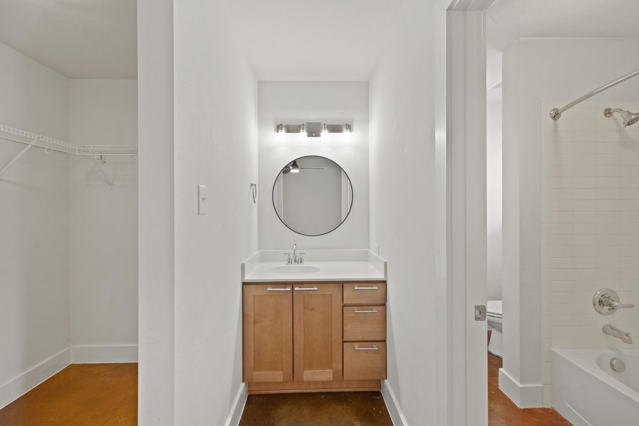 Bathroom vanity with a round mirror, beside an open closet and tiled bathtub.