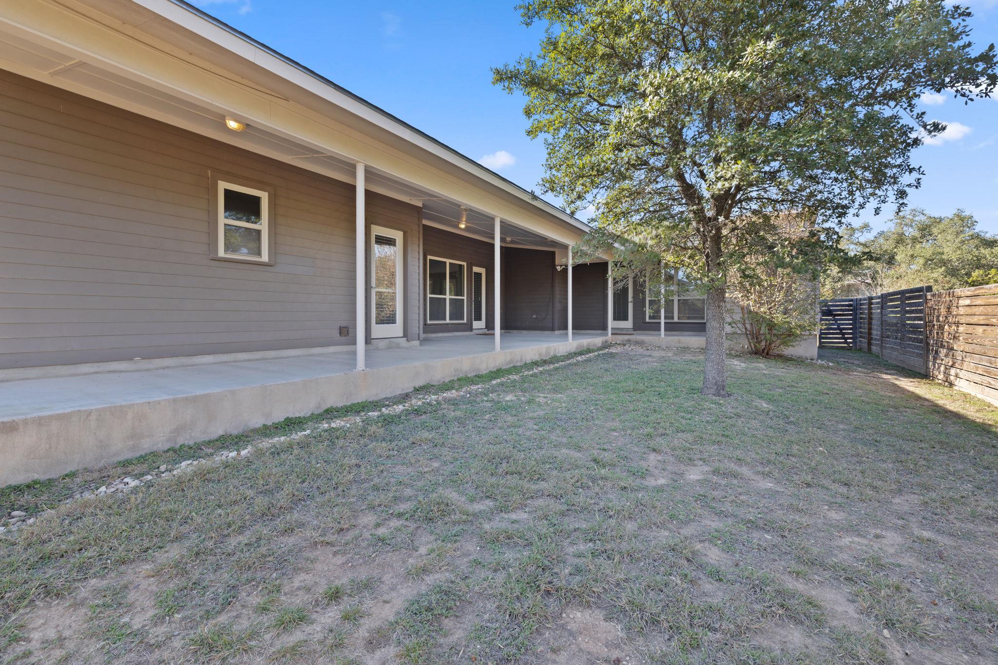 Home’s rear covered patio beside a large shade tree and fenced, patchy lawn.