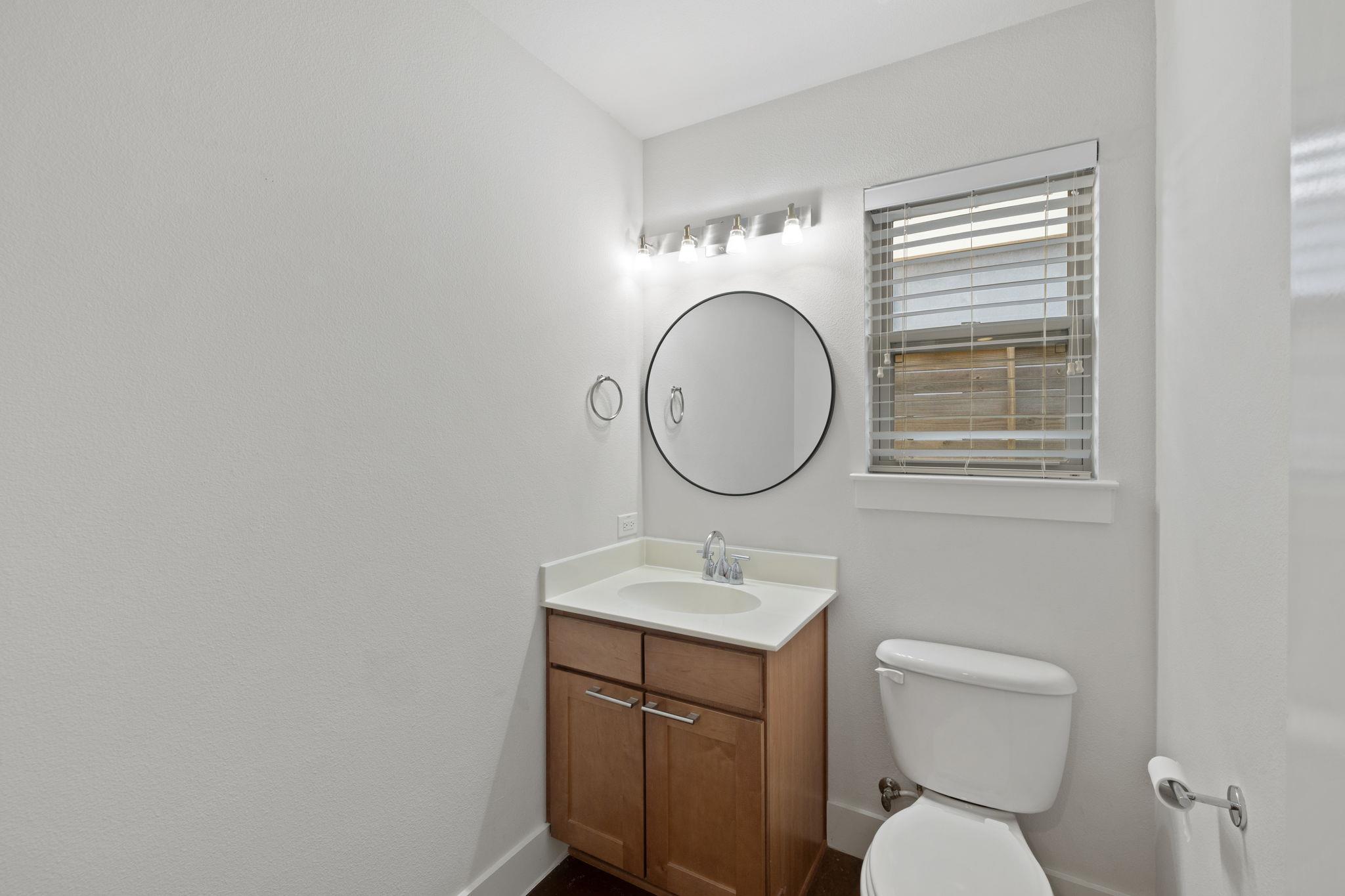 Bathroom with a wood vanity, round mirror, window with blinds, and toilet.