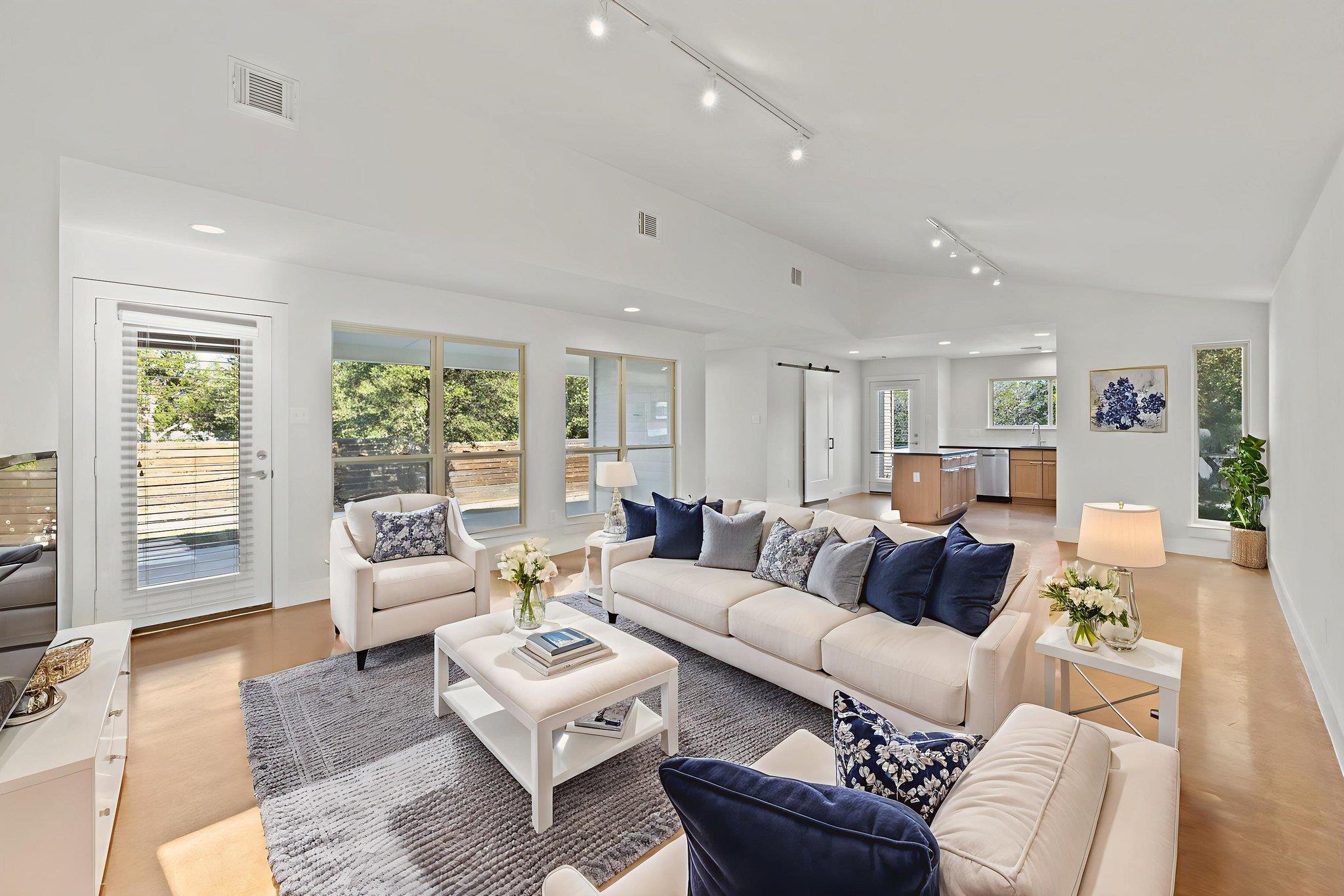 Staged living room with white sofas, navy cushions, large windows, and an open kitchen.