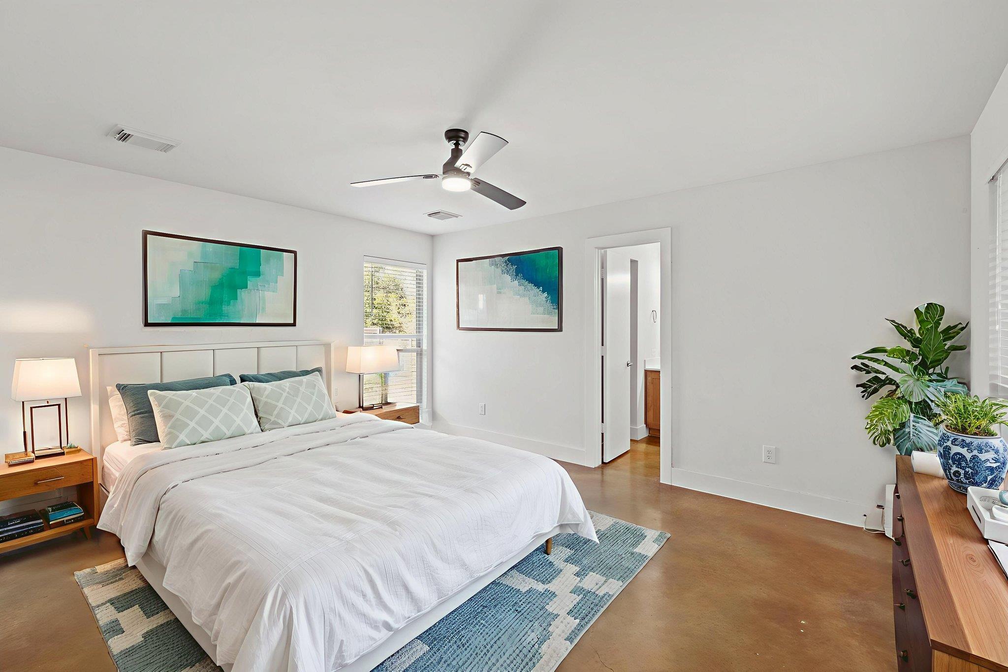 Bright staged bedroom with a white bed, turquoise wall art, and leafy plants beside a wooden dresser.