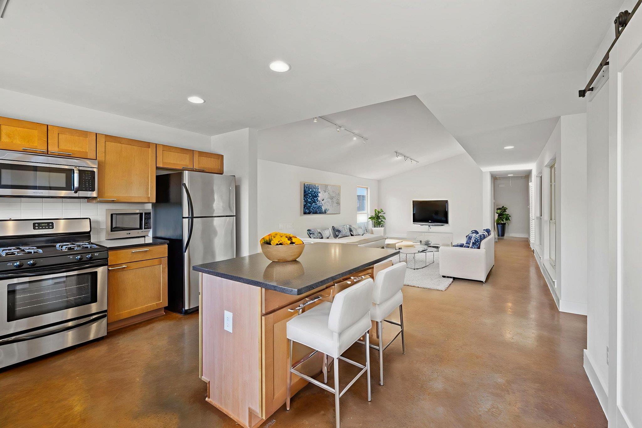 Open-plan kitchen and living room with wood cabinets, stainless appliances, and a dark island with white stools.