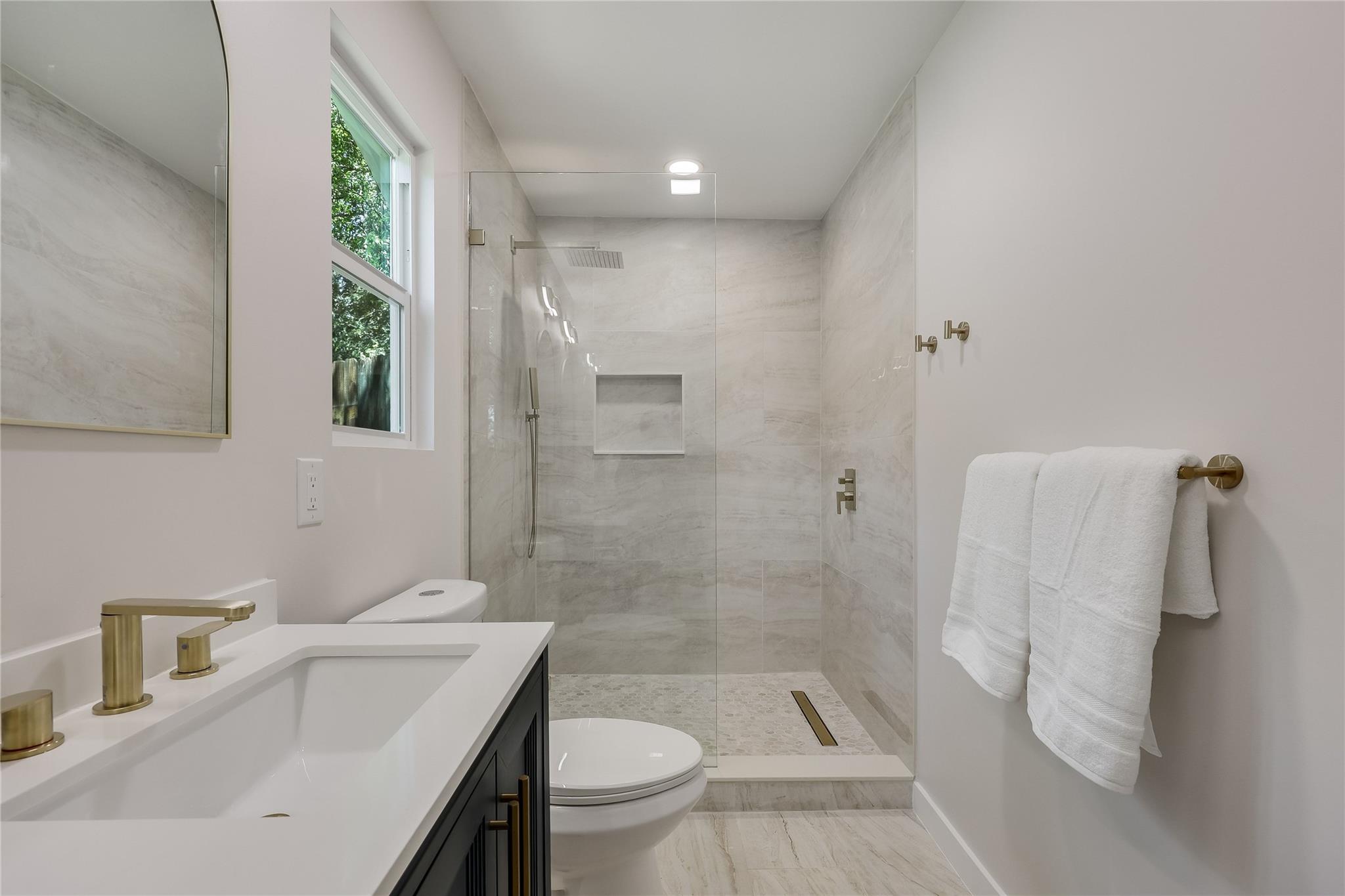 Bathroom with a glass walk-in shower, light tile, brass fixtures, and a window.
