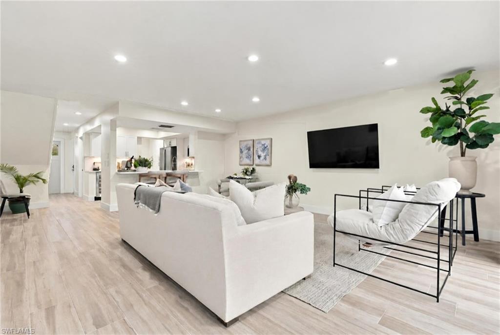 Staged living room with white seating, a wall-mounted television, and an open kitchen.