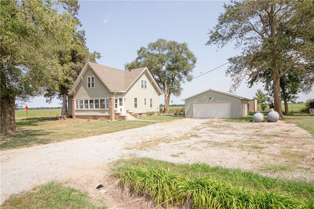 Light-colored two-story house beside a detached garage, with a gravel driveway and open fields.