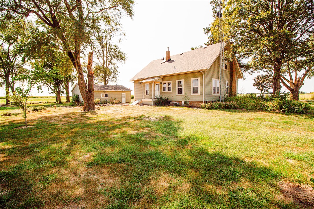 Tan farmhouse in a tree-shaded lawn, with a small outbuilding and open fields behind it.