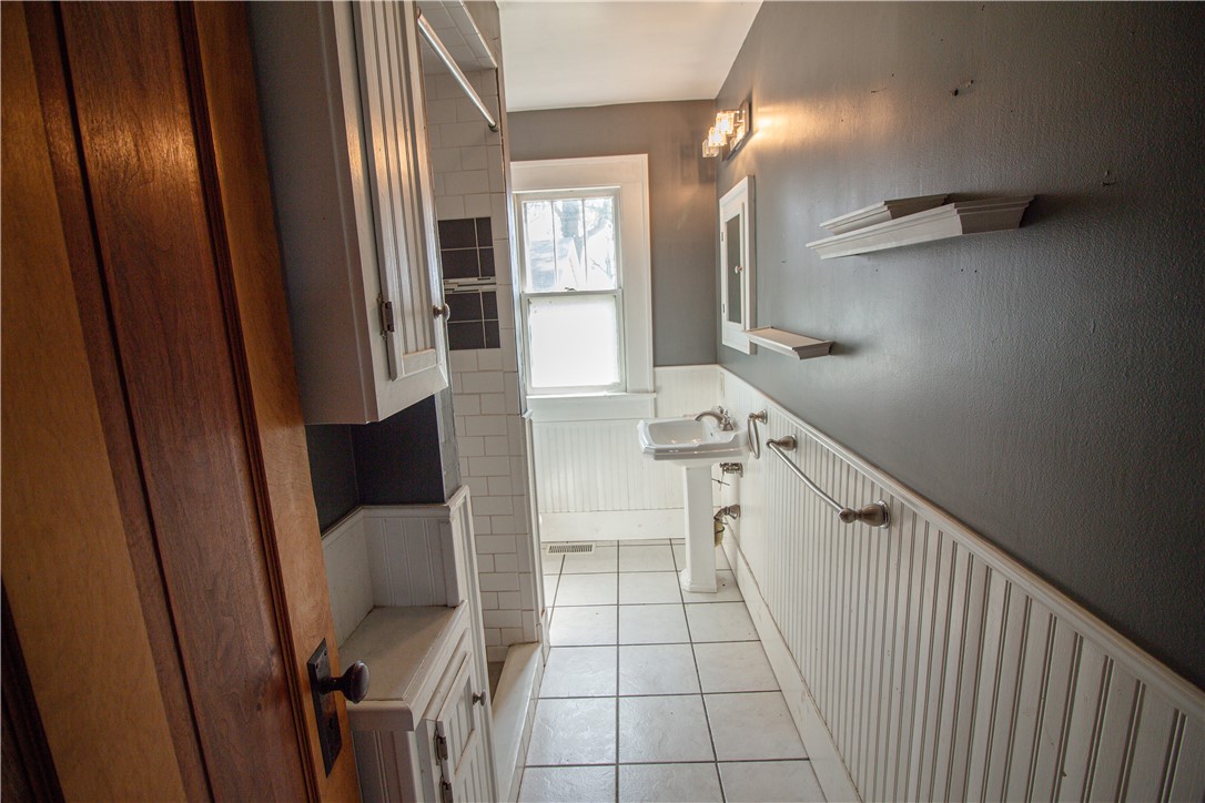 Narrow bathroom with a pedestal sink beneath a window and gray walls above white beadboard wainscoting.
