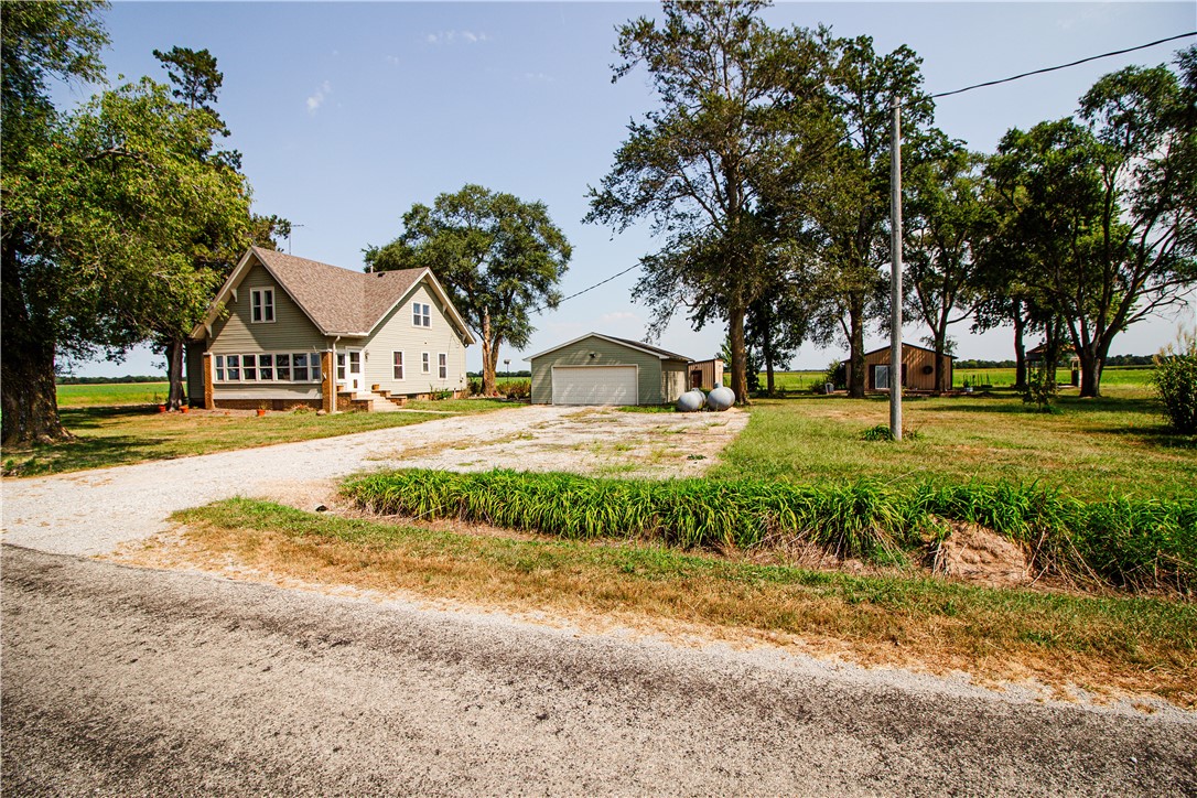 Rural home beside a gravel driveway, with a detached garage, outbuildings, and mature trees.