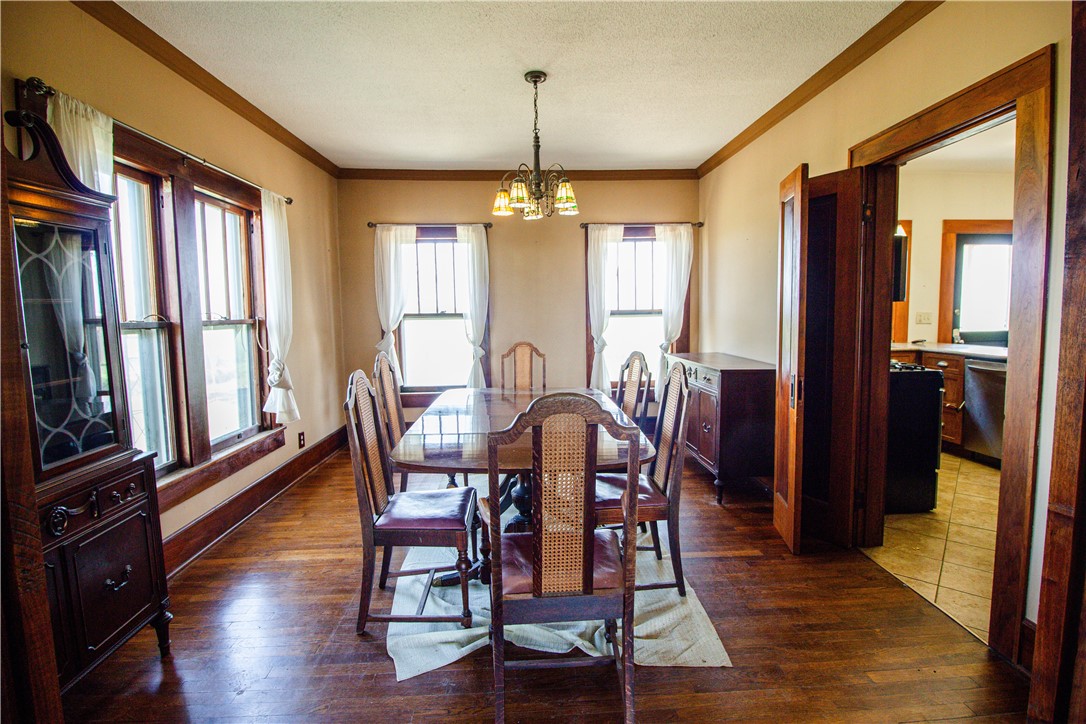 Dining room with a wooden table, hardwood floors, large curtained windows, and an adjoining kitchen.