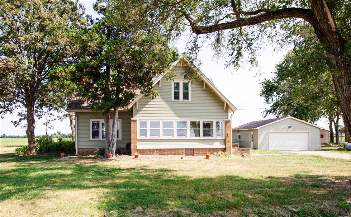 Sage-sided, brick-trimmed house with a detached garage, framed by mature trees and open fields.