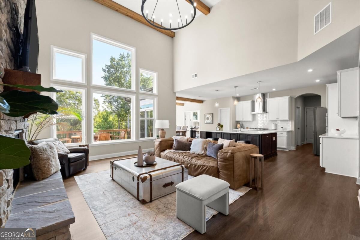 Furnished living room opening to a white kitchen, with a stone fireplace and tall windows.