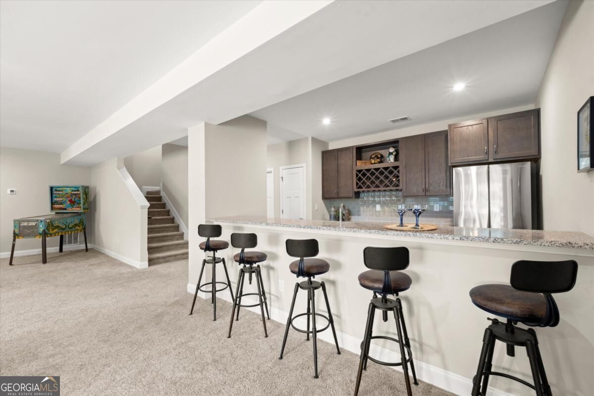 Finished basement bar with five stools, dark cabinetry, stainless-steel refrigerator, and carpeted recreation area.