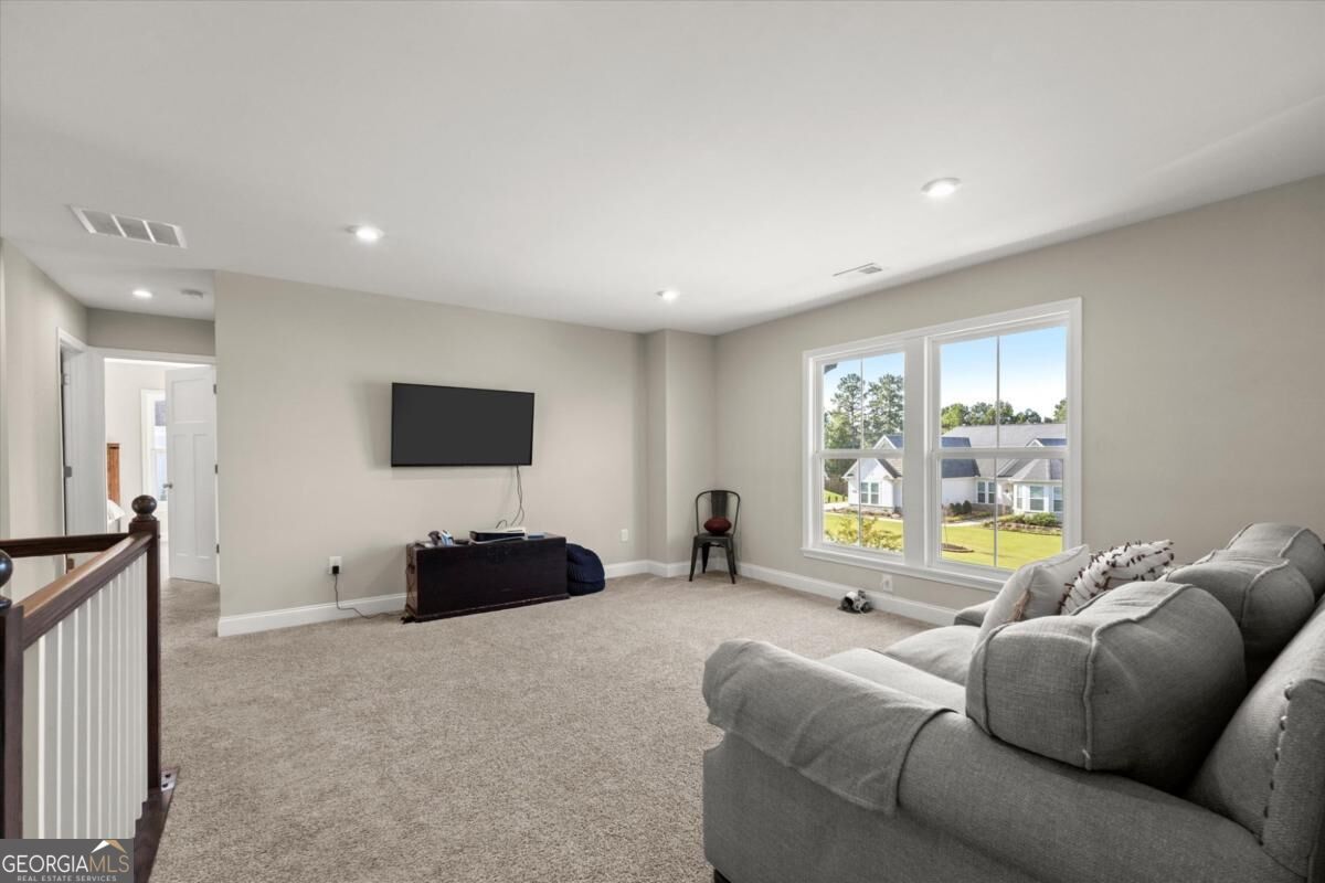 Carpeted living area with a gray sectional, wall-mounted TV, and windows overlooking neighboring homes.